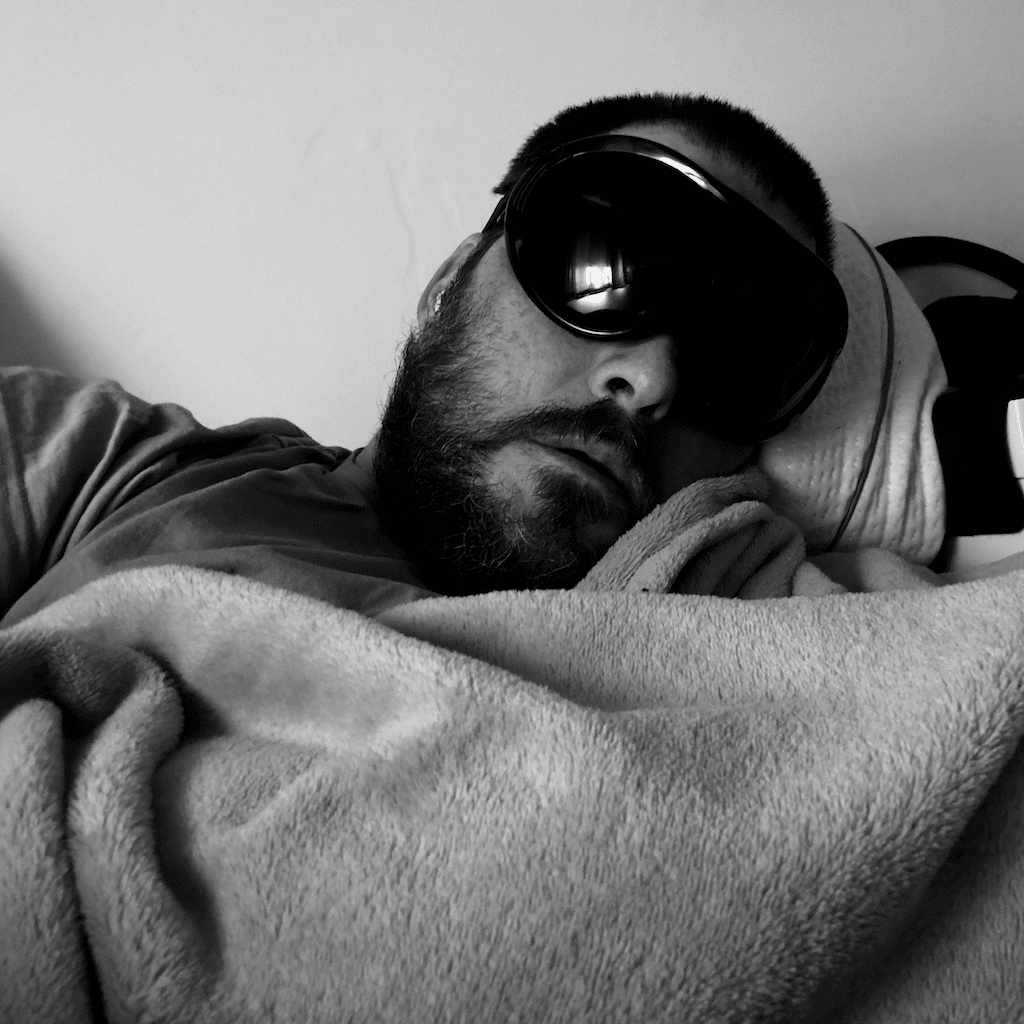 Taken in February 2026 wearing a heavy duty welding goggles to block out light and resting with a large blanket and pillow in a seated position. A moody self portrait in monochrome with the subject slightly out of focus.
